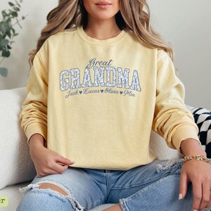 Pu&ograve; includere: Una felpa giallo chiaro con la scritta "Great Grandma" in un motivo floreale. Sotto, sono visualizzati i nomi Jack, Lucas, Olivia e Mia. La felpa &egrave; a maniche lunghe.