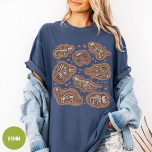 May include: A navy blue t-shirt featuring a graphic print of dinosaur skeletons in various poses. The skeletons are white and are surrounded by brown outlines. The shirt has a relaxed fit and a crew neck.