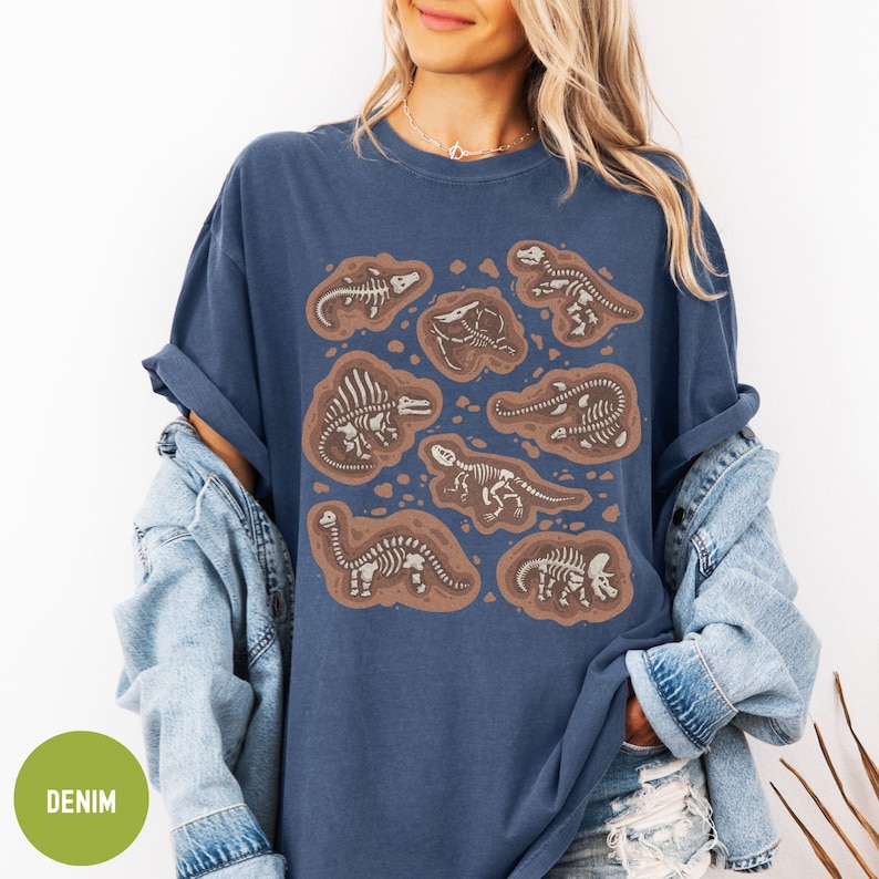 May include: A navy blue t-shirt featuring a graphic print of dinosaur skeletons in various poses. The skeletons are white and are surrounded by brown outlines. The shirt has a relaxed fit and a crew neck.