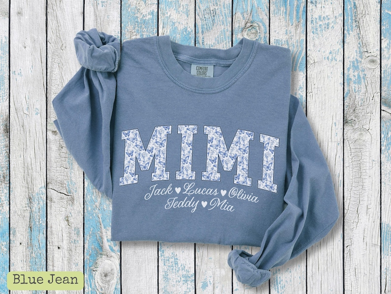 May include: A blue jean-coloured sweatshirt featuring the word "MIMI" in large, floral-patterned letters. Below, names are listed: Jack, Lucas, Olivia, Teddy, and Mia. The sweatshirt is folded on a light blue and white wooden surface.