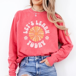 May include: A coral-colored long-sleeve sweatshirt with the text "LET'S LEARN TODAY" in a retro font. The design includes a sun graphic and small flower accents. The sweatshirt is paired with blue denim jeans.