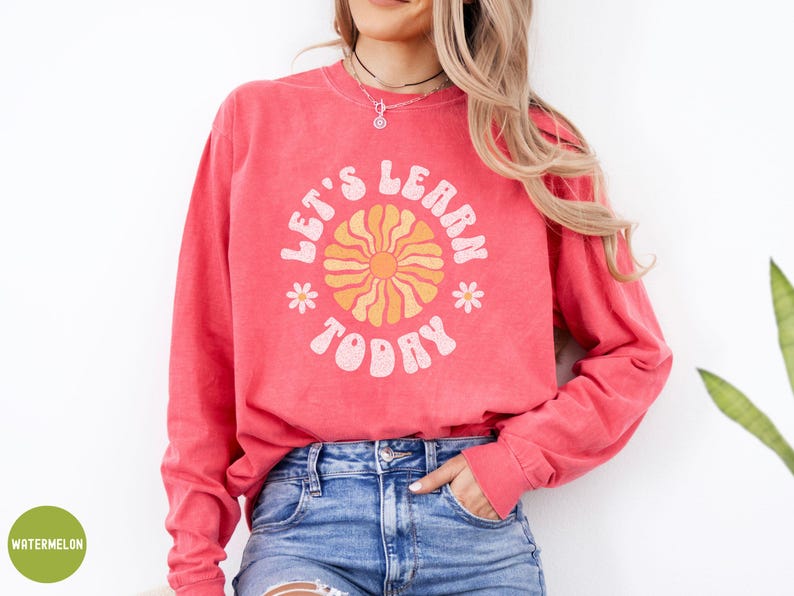 May include: A coral-colored long-sleeve sweatshirt with the text "LET'S LEARN TODAY" in a retro font. The design includes a sun graphic and small flower accents. The sweatshirt is paired with blue denim jeans.