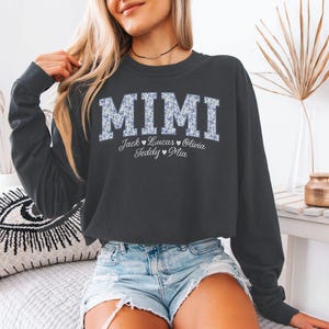 May include: A black crew neck sweatshirt featuring the word "MIMI" in a floral pattern. Underneath are the names Jack, Lucas, Olivia, Teddy, and Mia. The sweatshirt is worn with denim shorts.