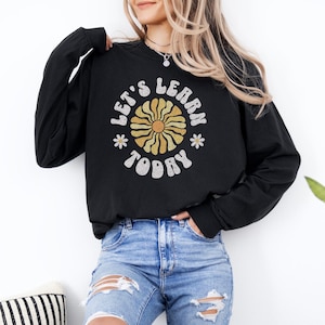 May include: Black long-sleeve sweatshirt with the text "LET'S LEARN TODAY" in a retro font. The design includes a yellow and gold flower graphic. The sweatshirt is worn with blue jeans.