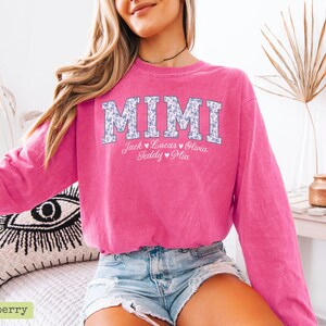 May include: A vibrant pink sweatshirt featuring the word "MIMI" in a floral pattern. Below the word are the names Jack, Lucas, Olivia, Teddy, and Mia. The sweatshirt is paired with denim shorts. The image is well-lit.