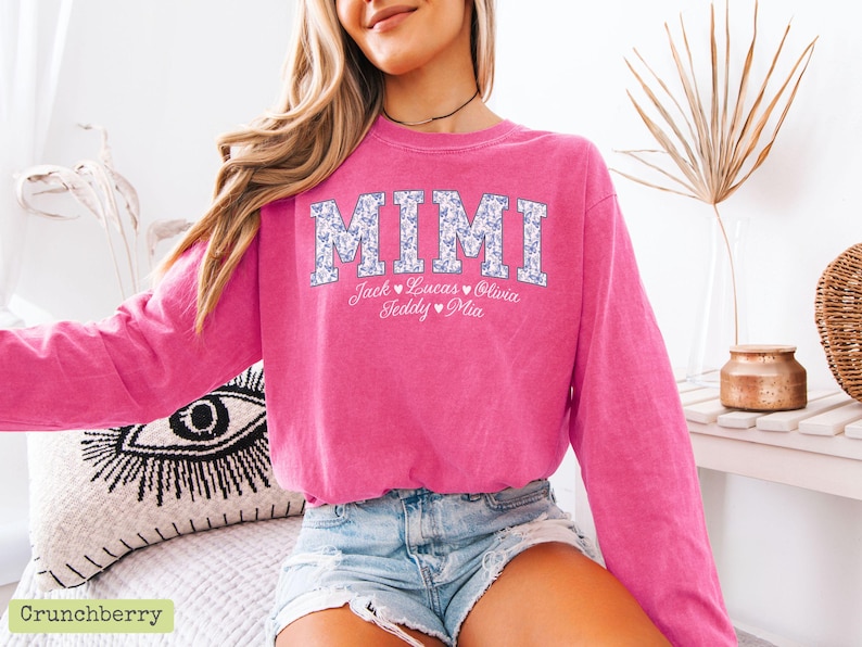 May include: A vibrant pink sweatshirt featuring the word "MIMI" in a floral pattern. Below the word are the names Jack, Lucas, Olivia, Teddy, and Mia. The sweatshirt is paired with denim shorts. The image is well-lit.