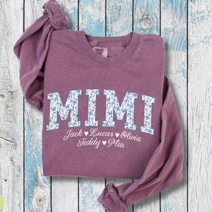 May include: Berry-coloured sweatshirt featuring the word "MIMI" in large, white letters with a blue floral pattern. Below "MIMI" are the names Jack, Lucas, Olivia, Teddy, and Mia. The sweatshirt is folded on a light blue and white wooden surface.