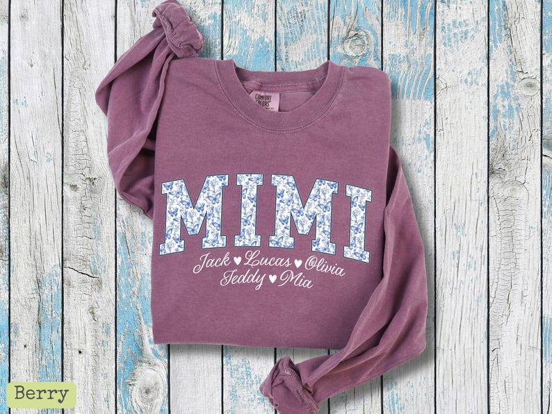 May include: Berry-coloured sweatshirt featuring the word "MIMI" in large, white letters with a blue floral pattern. Below "MIMI" are the names Jack, Lucas, Olivia, Teddy, and Mia. The sweatshirt is folded on a light blue and white wooden surface.