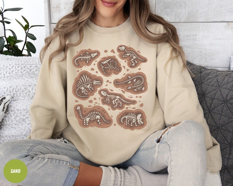 May include: A beige sweatshirt displaying several brown dinosaur skeleton illustrations. The design showcases different dinosaur species within brown outlines. The word "SAND" is in a green circle. The overall theme is prehistoric.