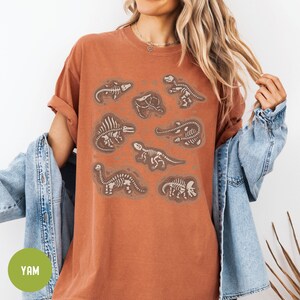 May include: A rust-coloured t-shirt with a graphic print of multiple dinosaur skeletons in various poses. The skeletons are white and are printed on a rust-coloured background.