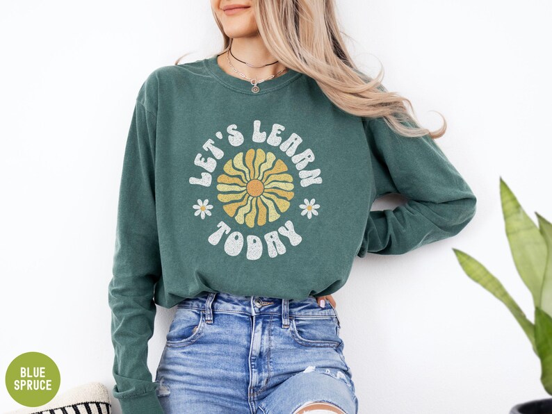 May include: Green long-sleeve sweatshirt with the text "LET'S LEARN TODAY" in white and a yellow and orange flower design. The sweatshirt is paired with blue jeans.