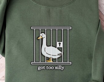May include: An olive green crewneck sweatshirt featuring an embroidered graphic of a white goose with an orange beak and feet standing inside a white cage. Below the cage, the text "got too silly" is embroidered in a white, lowercase font.