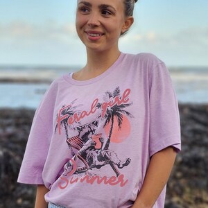 May include: A woman wearing a light pink t-shirt with the text "Feral Girl Summer" in pink and black. She is wearing light blue denim shorts and has her hair pulled back in two buns.
