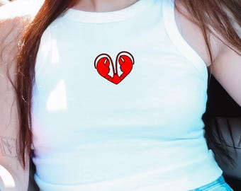 Puede incluir: Camiseta sin mangas blanca acanalada con un diseño gráfico en forma de corazón rojo. El diseño presenta dos figuras estilizadas dentro del corazón. La camiseta se combina con vaqueros de mezclilla azul claro. La imagen está tomada con luz natural.