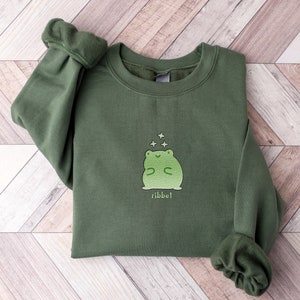May include: A dark green sweatshirt with a light green frog graphic and the word "ribbit" printed below. The frog is surrounded by small white stars.