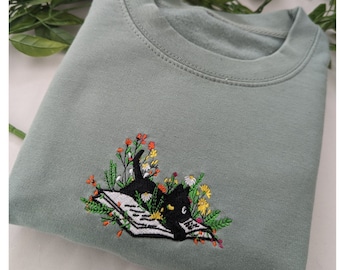 May include: A sage green sweatshirt with an embroidered design of a black cat reading a book surrounded by flowers.