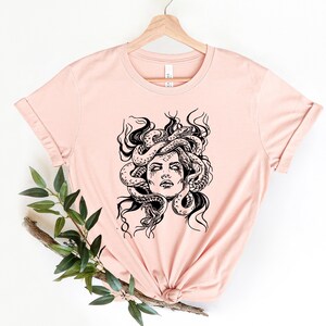 May include: A light pink t-shirt with a black and white illustration of Medusa, a Greek mythological figure with snakes for hair.