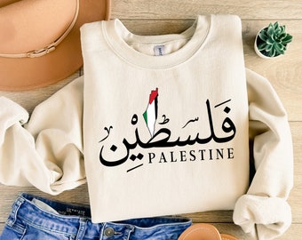 May include: Cream-colored sweatshirt with the word "Palestine" in Arabic script and English text. The design includes a map of Palestine with the colors of the Palestinian flag. The sweatshirt is paired with blue jeans, a hat, and sandals.