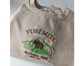 May include: A beige sweatshirt with an embroidered Yosemite National Park logo. The logo features a brown bear, green trees, and a yellow sun.