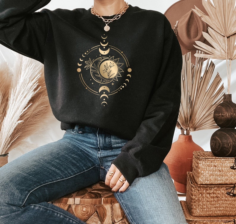 May include: Black sweatshirt featuring a gold moon and floral design. The design depicts a crescent moon with a floral stem and leaves inside the moon. The moon is encircled by a circle of smaller moons and stars.