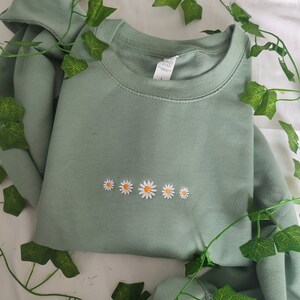 Pu&ograve; includere: Felpa verde salvia con una fila di cinque margherite bianche ricamate.