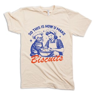 May include: Cream-colored t-shirt featuring a retro illustration of a woman and a cat wearing chef hats, mixing ingredients in a bowl. The text "SO THIS IS HOW U MAKE Biscuits" is printed in blue and red.