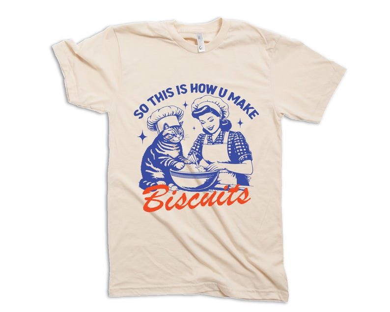 May include: Cream-colored t-shirt featuring a retro illustration of a woman and a cat wearing chef hats, mixing ingredients in a bowl. The text "SO THIS IS HOW U MAKE Biscuits" is printed in blue and red.
