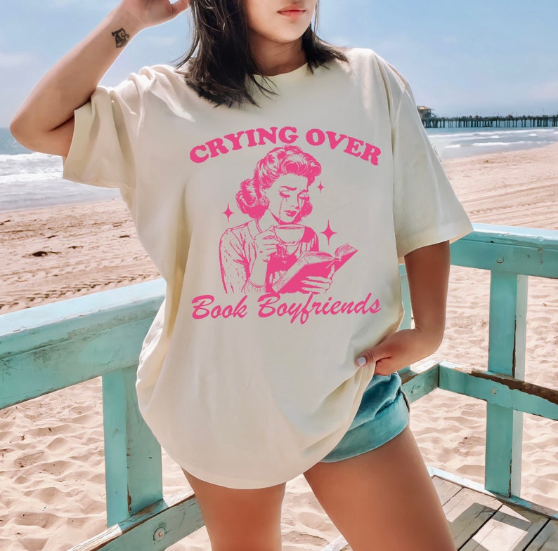 May include: A white t-shirt with a pink graphic of a woman reading a book and crying. The text on the shirt reads "Crying Over Book Boyfriends".