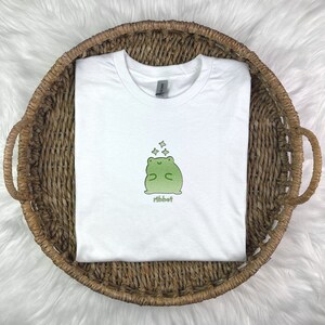 May include: White t-shirt with a green frog illustration and the word "ribbit" printed below. The frog is surrounded by small white stars.