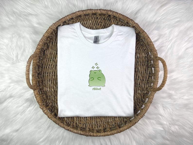 May include: White t-shirt with a green frog illustration and the word "ribbit" printed below. The frog is surrounded by small white stars.