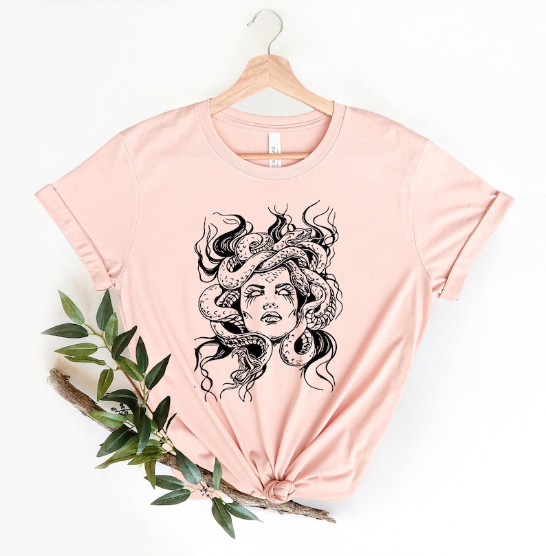 May include: A light pink t-shirt with a black and white illustration of Medusa, a Greek mythological figure with snakes for hair.