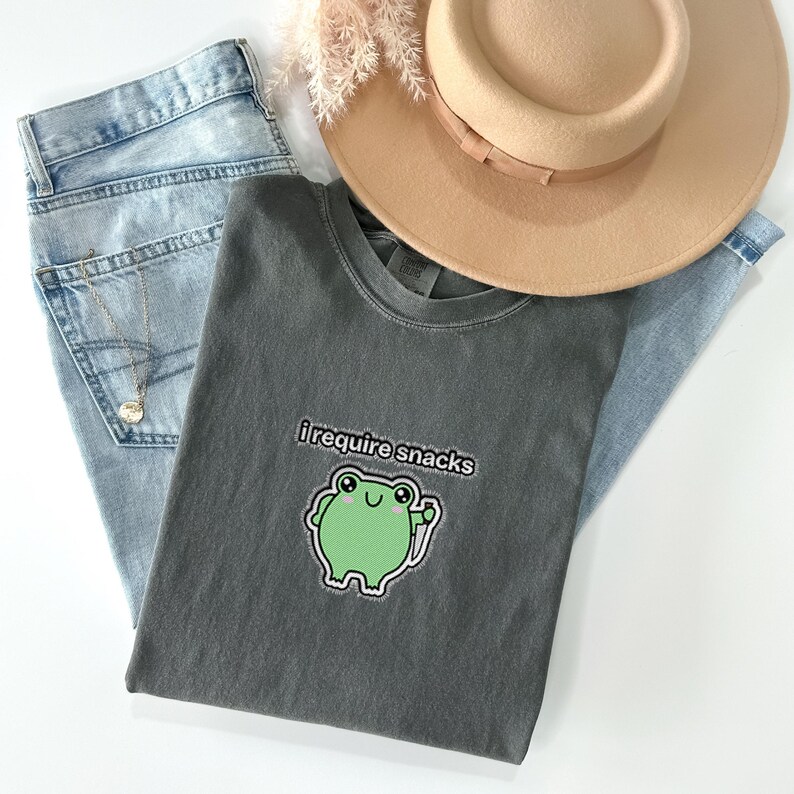 May include: A casual fashion ensemble featuring a gray t-shirt with the text "i require snacks" and a green frog graphic. The outfit includes light blue jeans, a tan fedora hat, and a gold necklace. The items are arranged on a white surface.