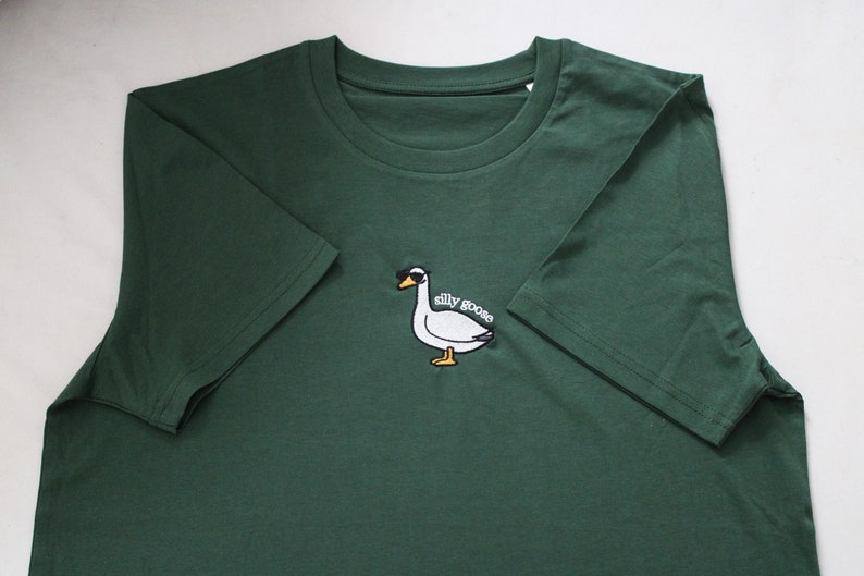 May include: A dark green t-shirt featuring a white embroidered goose wearing sunglasses and the text "silly goose".