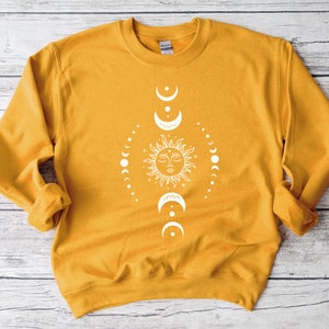 May include: A mustard yellow sweatshirt with a white sun and moon design. The sun has a face and is surrounded by a circle of moons in different phases.
