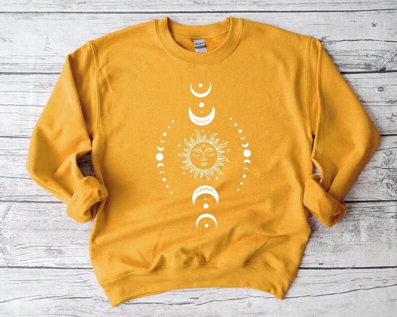 May include: A mustard yellow sweatshirt with a white sun and moon design. The sun has a face and is surrounded by a circle of moons in different phases.