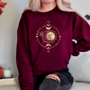 Peut inclure: Un sweat-shirt bordeaux avec un motif soleil et lune dor&eacute; repr&eacute;sentant un croissant de lune, une pleine lune et un soleil avec un motif floral au centre. Le motif est entour&eacute; d'un cercle de petites lunes.