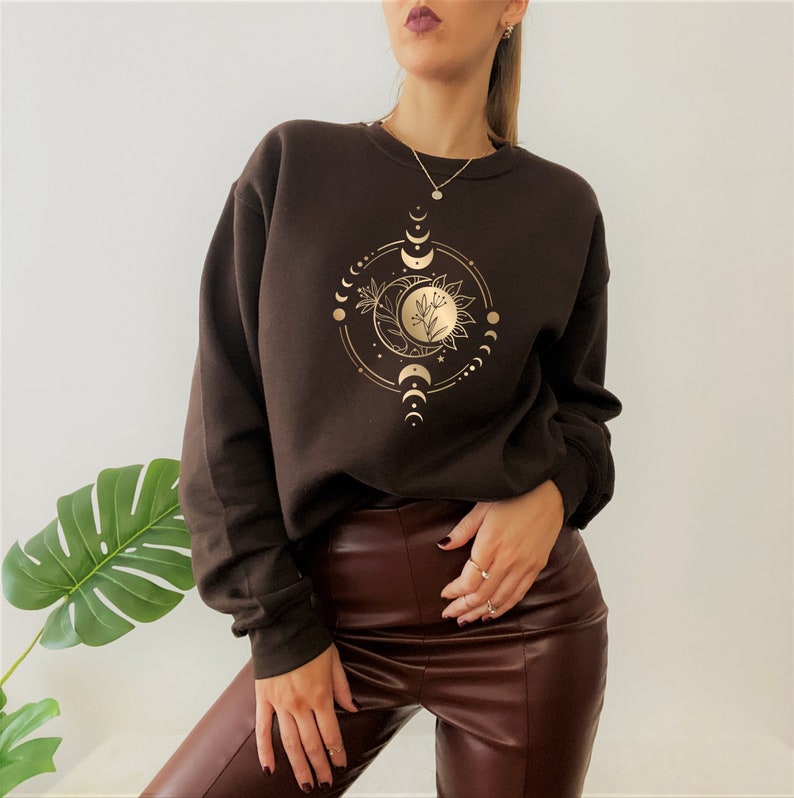 Peut inclure: Un sweat-shirt marron avec un motif dor&eacute; d'une lune, d'&eacute;toiles et d'une fleur. Le motif est centr&eacute; sur la poitrine du sweat-shirt.