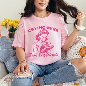 May include: A pink t-shirt with the text "Crying Over Book Boyfriends" and a retro illustration of a woman reading a book and drinking coffee.