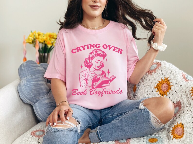 May include: A pink t-shirt with the text "Crying Over Book Boyfriends" and a retro illustration of a woman reading a book and drinking coffee.