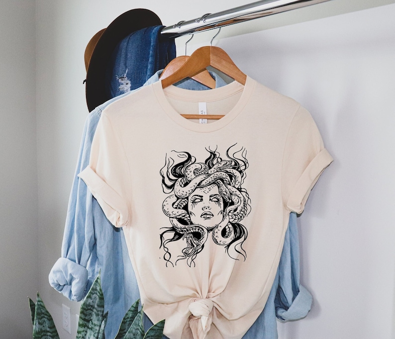 May include: A beige t-shirt with a black and white graphic print of Medusa, a mythical Greek figure with snakes for hair.