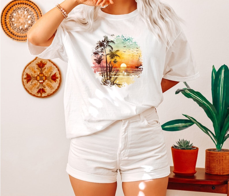 May include: White t-shirt featuring a colourful graphic print of palm trees and a sunset on the beach.