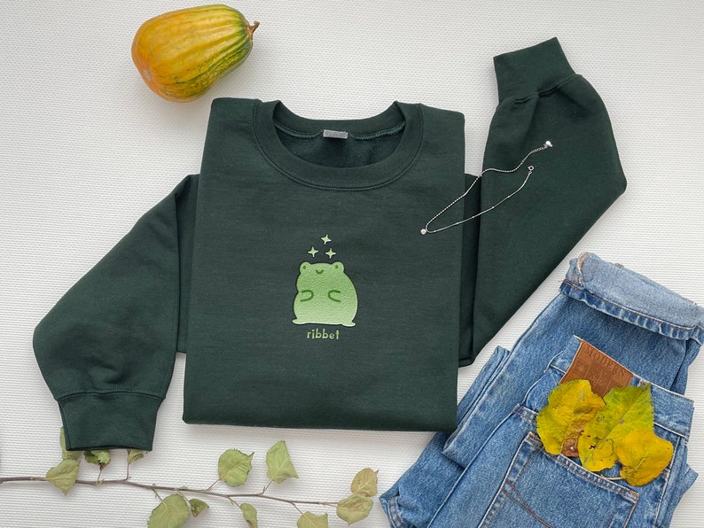 May include: A dark green sweatshirt with a light green frog graphic and the word "ribbit" printed below. The frog is surrounded by small white stars.