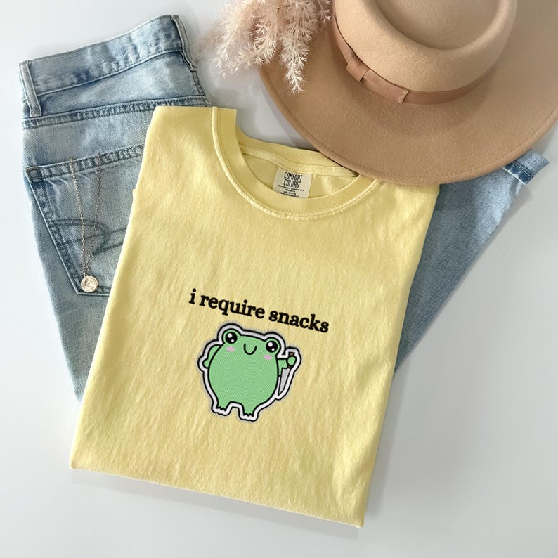 May include: A light yellow t-shirt with the phrase "i require snacks" and a green frog graphic. The shirt is paired with light blue jeans, a tan hat, and a gold necklace.