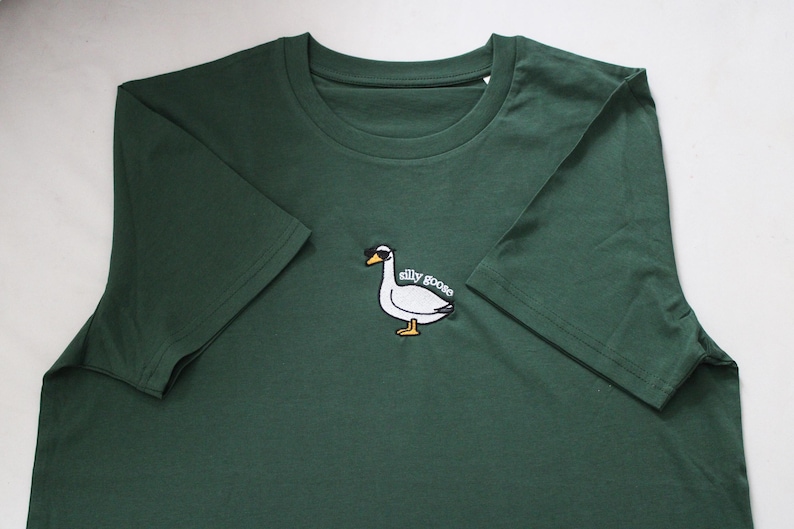 May include: A dark green t-shirt with a white embroidered goose wearing sunglasses and the text "silly goose".
