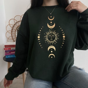 May include: A dark green sweatshirt with a gold sun and moon design. The sun has a face and is surrounded by a circle of smaller moons.