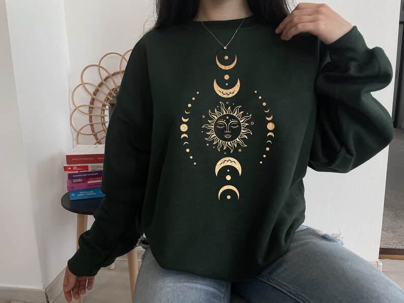 May include: A dark green sweatshirt with a gold sun and moon design. The sun has a face and is surrounded by a circle of smaller moons.