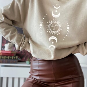 May include: A beige sweatshirt with a white sun and moon design. The sun has a face and is surrounded by a circle of moons.