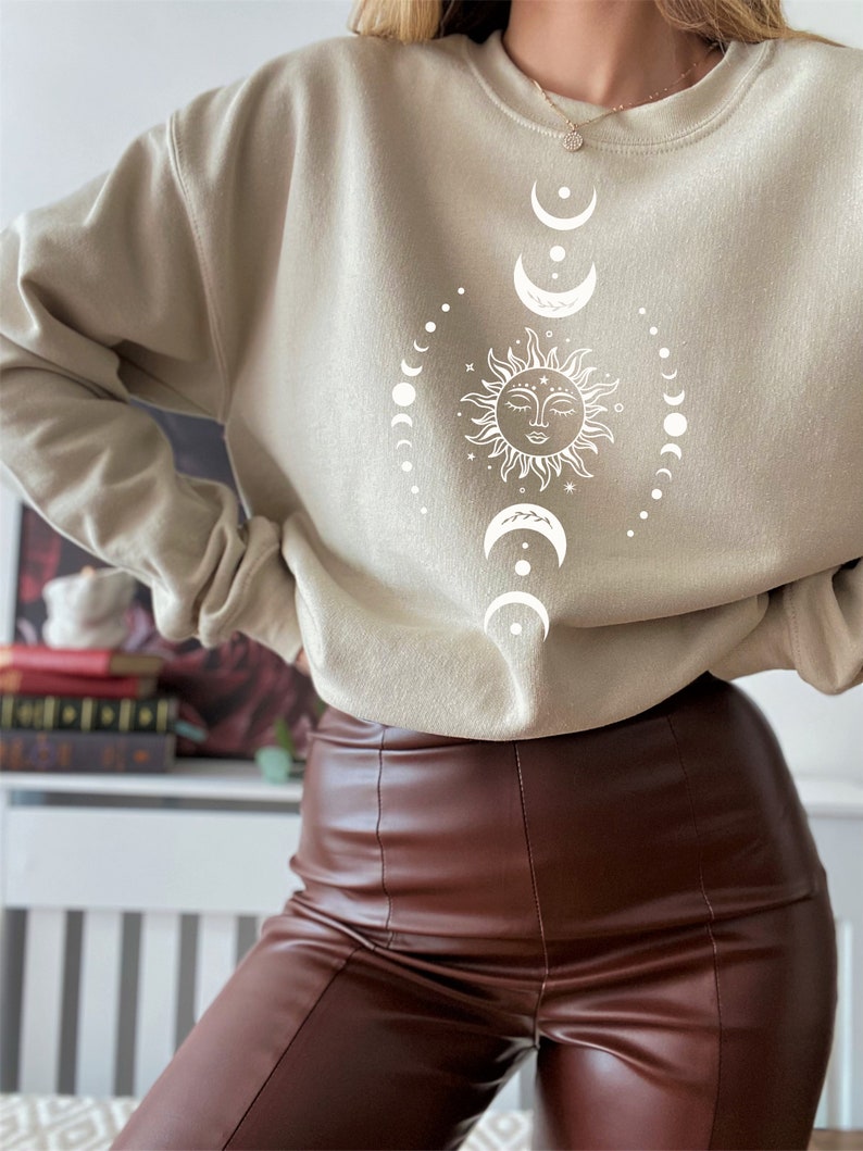 May include: A beige sweatshirt with a white sun and moon design. The sun has a face and is surrounded by a circle of moons.