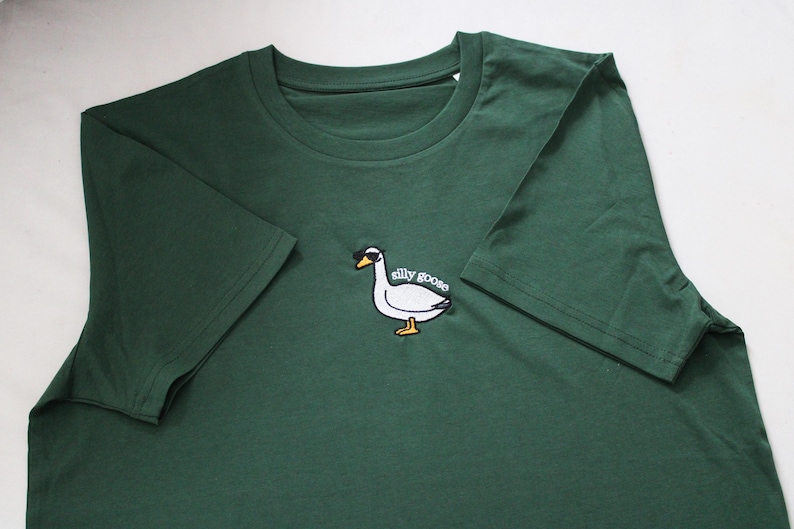 May include: A dark green t-shirt with an embroidered white goose wearing sunglasses and the text "silly goose".