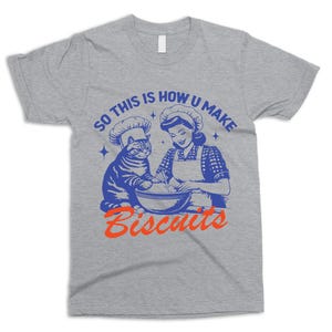 May include: A heather gray t-shirt featuring a blue and orange graphic. The graphic depicts a cat and a woman wearing chef hats, mixing ingredients in a bowl. The text reads "SO THIS IS HOW U MAKE BISCUITS."